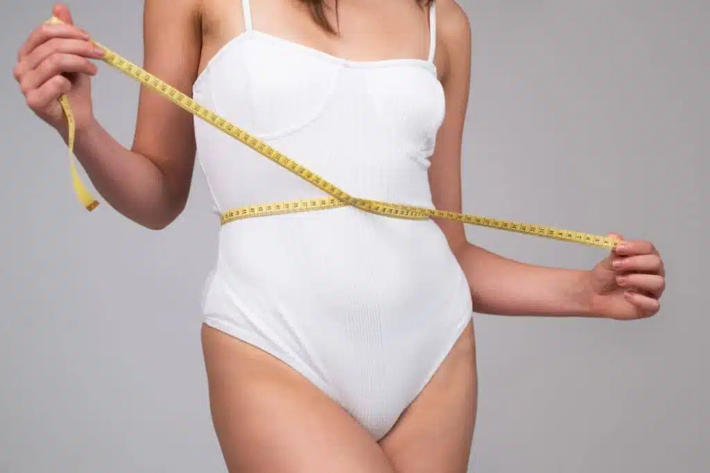 Woman measuring slim waistline in white bodysuit to show aesthetic and medical benefits of abdominoplasty.